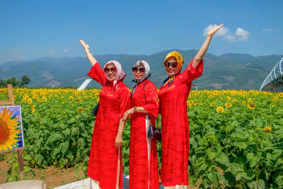 Ba phụ nữ trong tà áo dài đỏ nổi bật giữa cánh đồng hướng dương vàng óng dưới nền trời xanh, tạo nên khung hình rực rỡ, tràn sức sống